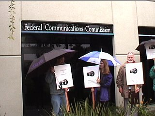 [Pic: protestors with signs & umbrellas outside FCC office]